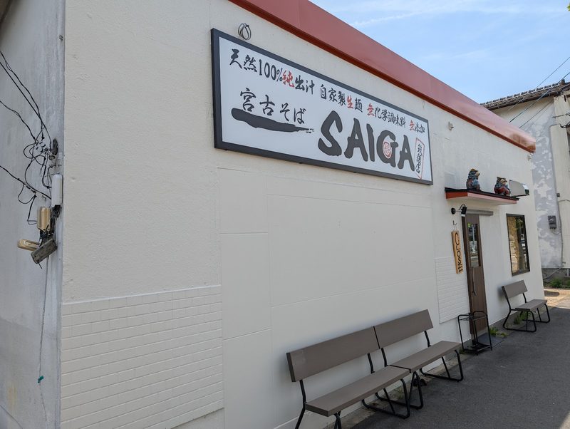 横から見た、白い壁の「SAIGA」の看板と店の前のベンチ